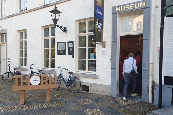 Der Eingang zum Museum Thorn befindet sich in einem weißen historischen Gebäude mit einer offenen Tür, durch die die Besucher eintreten. Vor dem Gebäude steht ein Holzschild mit der Aufschrift „THORN“ und einem Logo. An der Wand sind einige Fahrräder geparkt, und an der Fassade hängt eine Laterne.